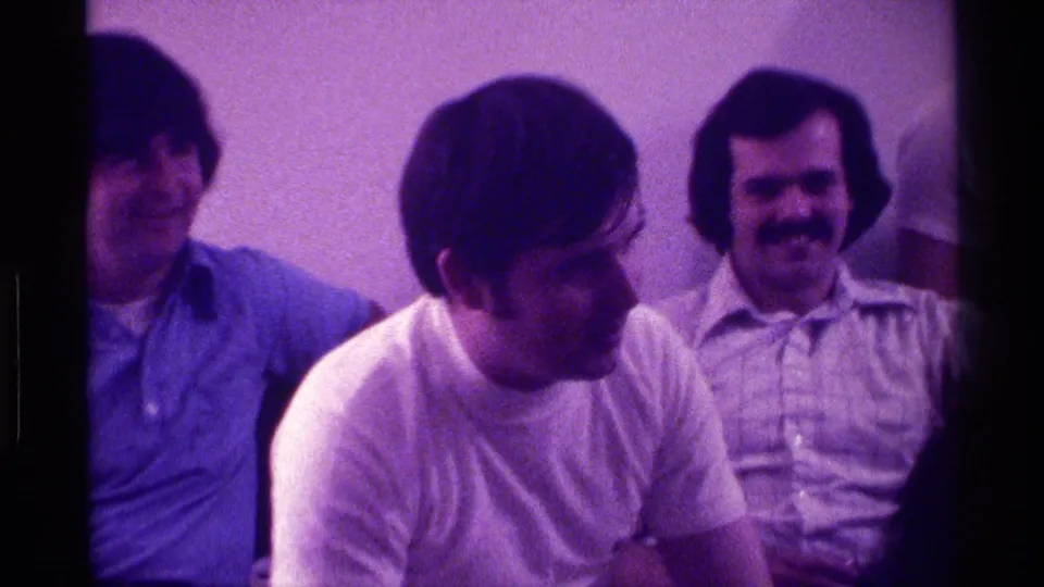 1976 Kentucky indoor home-movie still showing three adult men seated together, one speaking while a mustached man smiles.