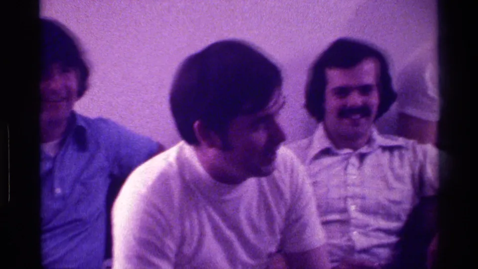 1976 Kentucky frame of a man in profile speaking during a casual indoor gathering, with two listeners behind him.