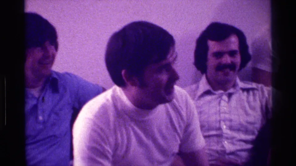 1976 Kentucky home-movie still of seated adult men sharing a light moment in a private indoor room.