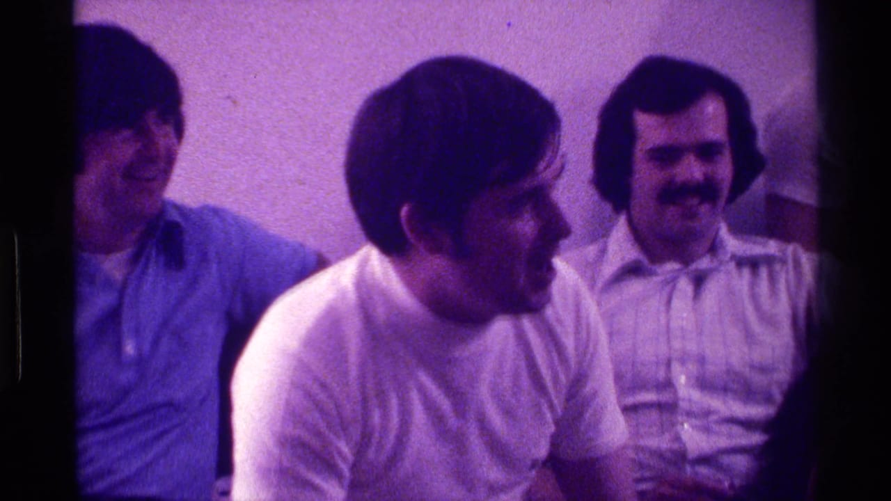 Three men sit indoors, laughing and watching someone off frame.