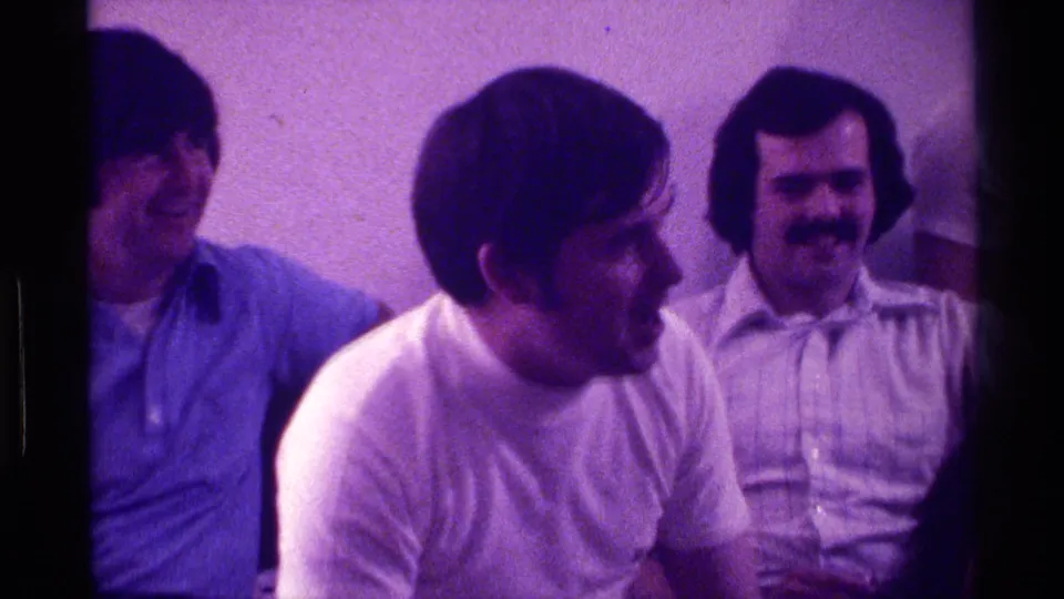 In supported 1976 Kentucky home-movie footage, three adult men sit against a plain wall while laughing and looking toward action off camera.
