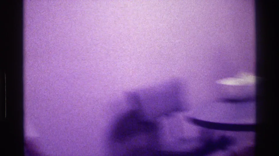 A heavily blurred figure passes through a plain private interior, with a small pale bowl or lamp-like object visible at the right edge.