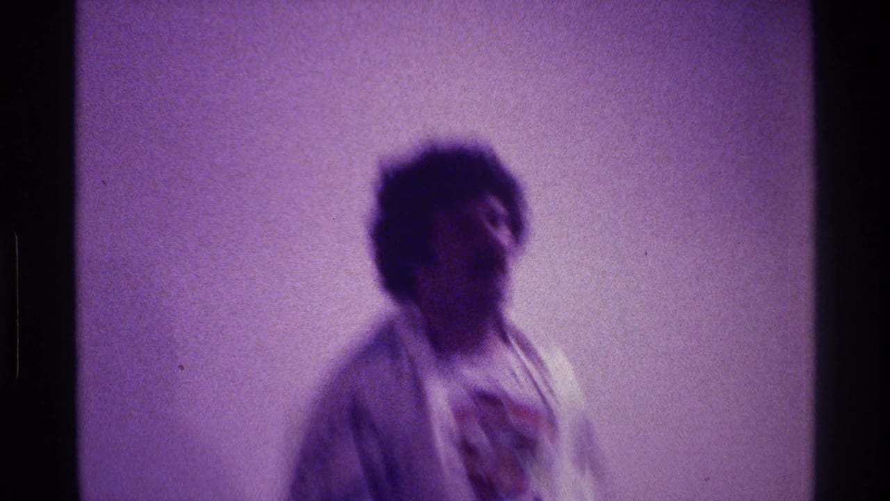 Blurred man in patterned fabric dancing against a plain interior wall.