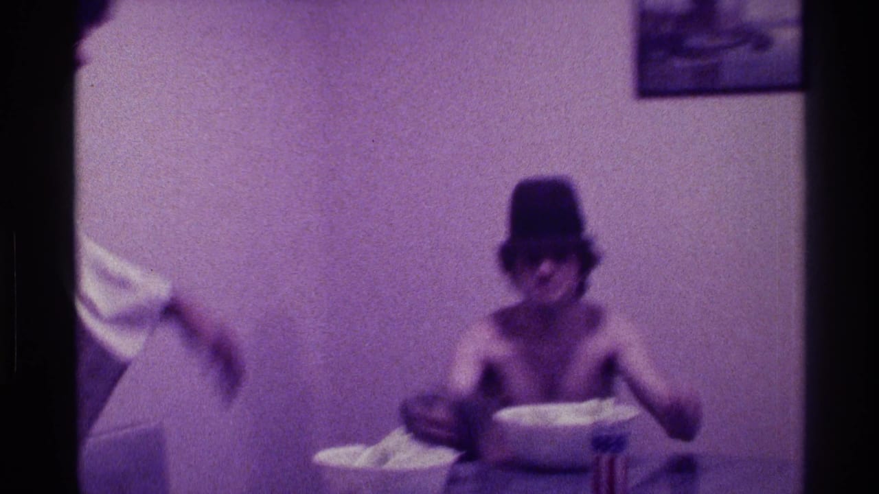 Purple-tinted 8mm frame of a shirtless man in a hat seated indoors with a bowl of popcorn on the table.