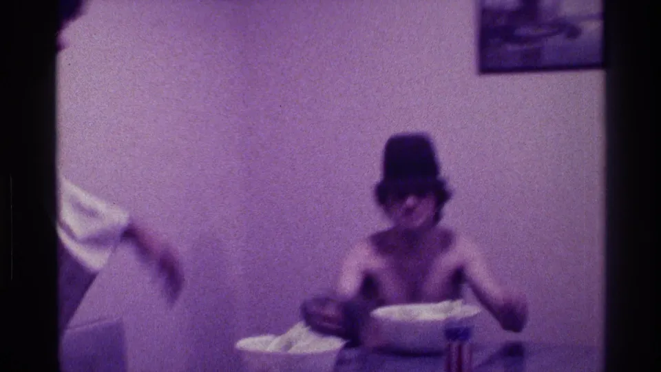 Purple-tinted 8mm frame of a shirtless man in a hat seated indoors with a bowl of popcorn on the table.