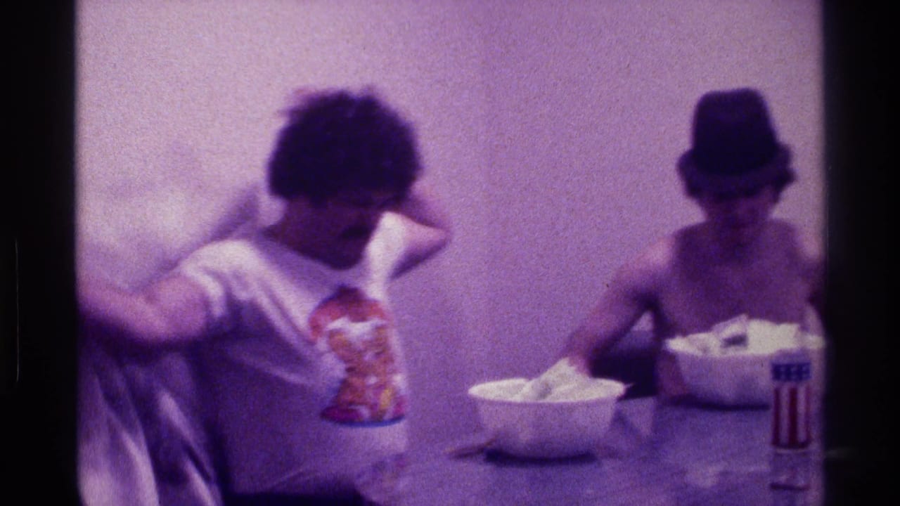 Two adult men sit at a table with bowls in a dim room.