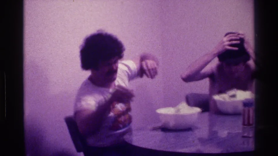 The seated man in a white graphic T-shirt gestures toward the table while the shirtless man beside him handles a dark hat.