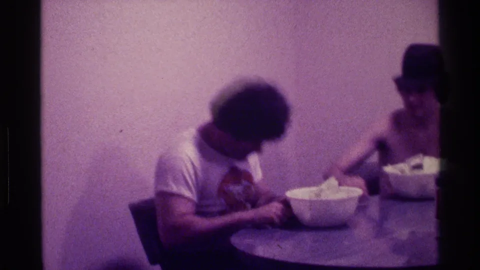 Two adult men in a plain indoor room lean toward bowls on a round table, with soft focus and a strong purple film cast.