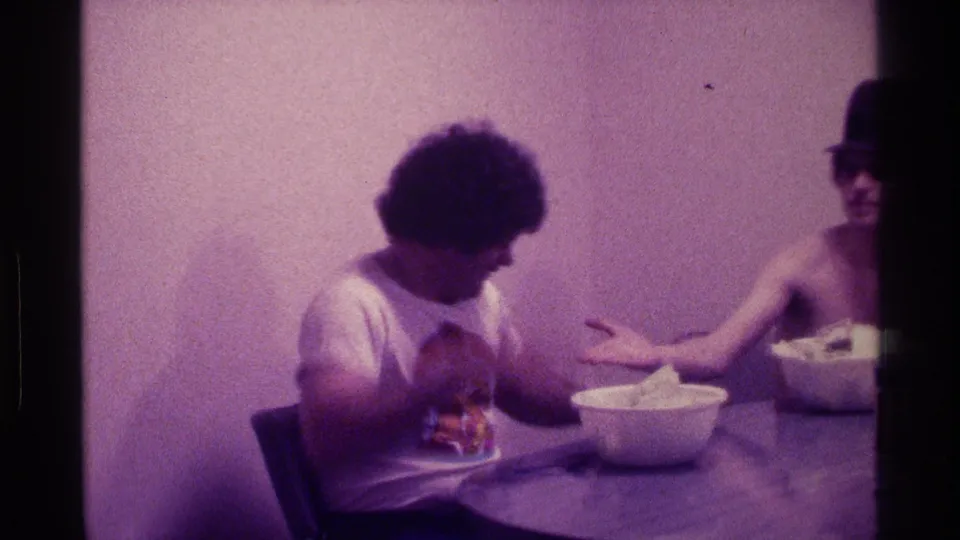 At the end of the clip, the man in the graphic T-shirt looks down as the shirtless man in a dark hat gestures beside a bowl.