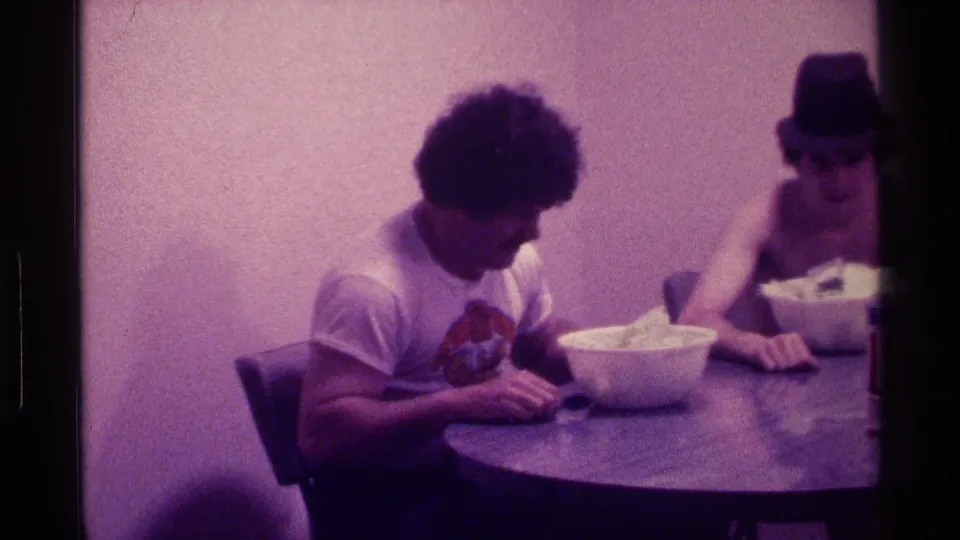 1976 Kentucky home-movie frame showing a young man in a graphic T-shirt looking down near a bowl while another young man in a black hat sits beside him.