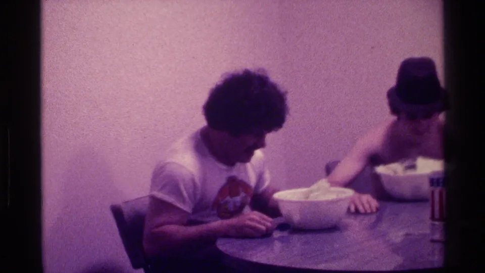 Purple-tinted 1976 Kentucky 8mm frame of two young men seated at a round table with white bowls and a small can near the edge.