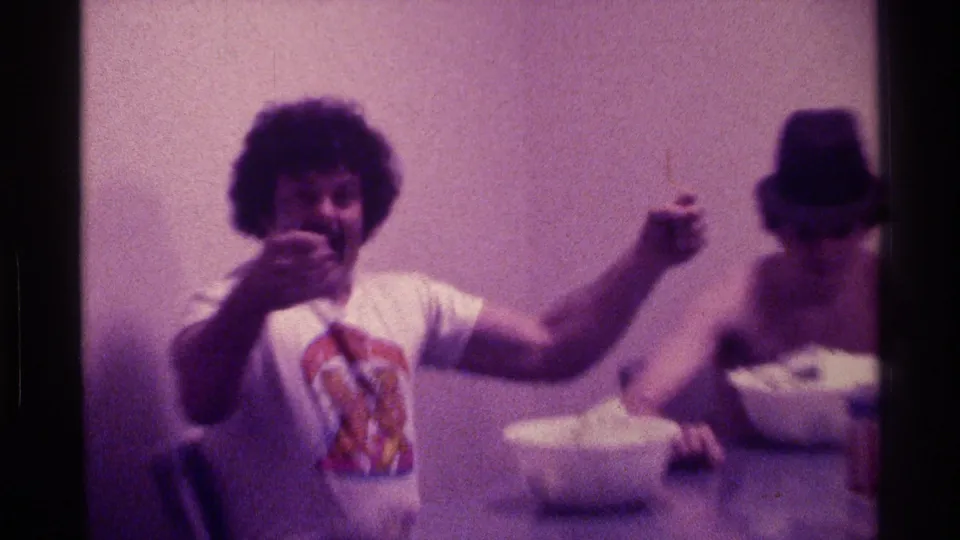 1976 Kentucky 8mm frame of a smiling young man in a graphic T-shirt gesturing toward the camera as another young man sits by a bowl.