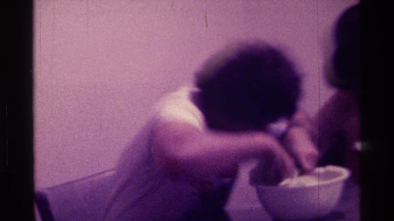 Person bends over a white bowl at a table while another person sits nearby.