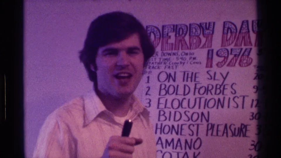 1976 Kentucky frame showing a man with a handheld microphone standing before a handwritten Derby Day board listing race entries.