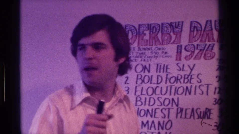1976 Kentucky home-movie frame of a man holding a microphone beside a handwritten Derby Day race board with visible horse names.