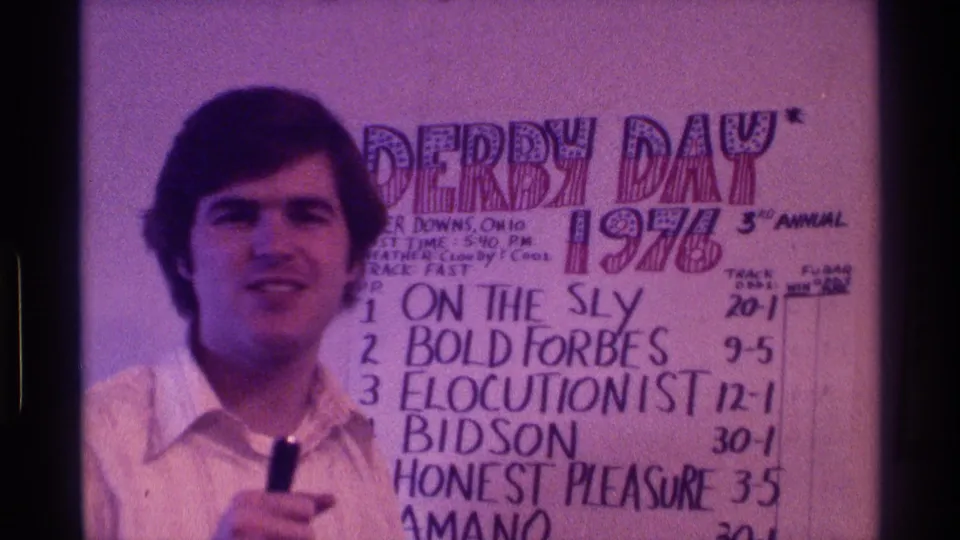 In 1976 Kentucky home-movie footage, a man in a white collared shirt faces the camera while holding a microphone beside a handwritten Derby Day poster.