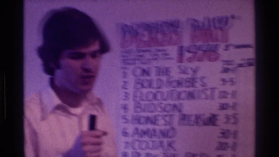 The man continues speaking into a handheld microphone beside the Derby Day 1976 poster, with the frame softened by motion blur and purple 8mm color shift.
