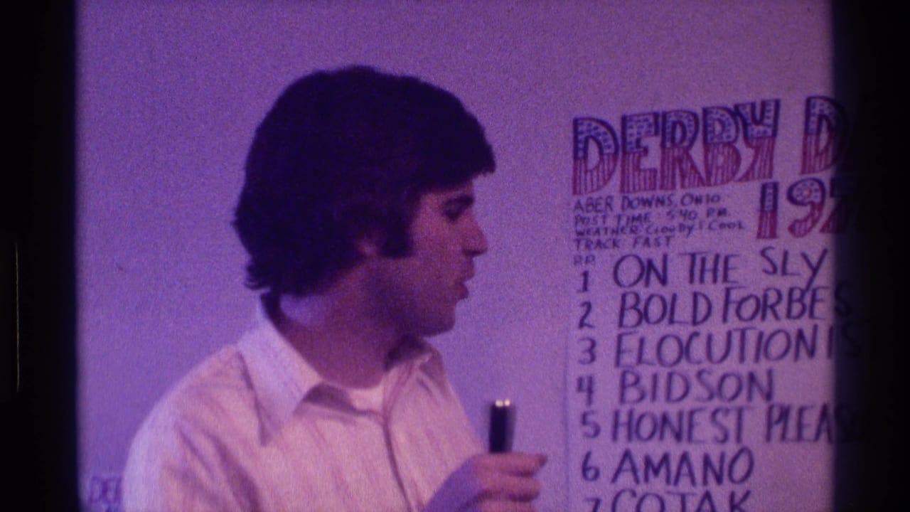Man in a white collared shirt holds a microphone beside a handwritten Derby Day board.