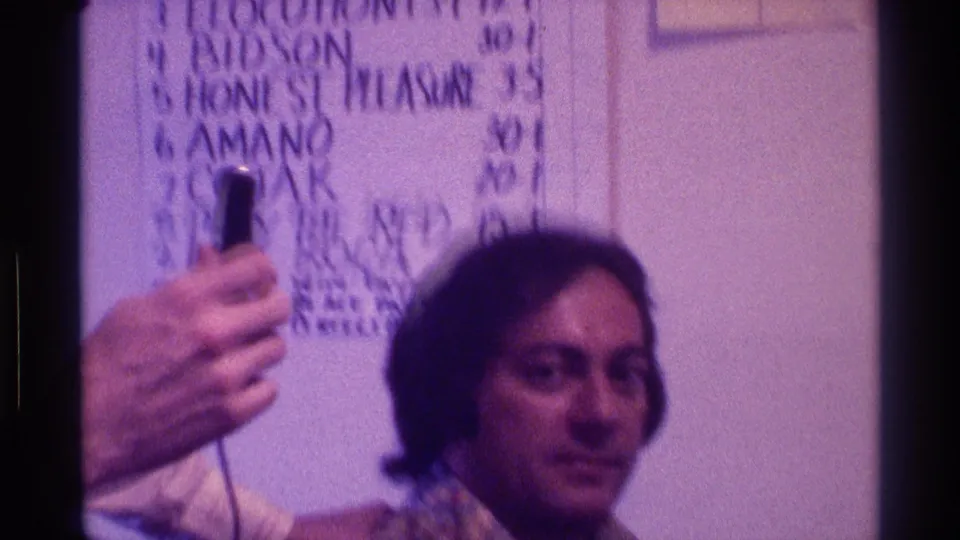 A seated man with longer hair looks toward the camera as the microphone and Derby Day board remain visible in the Kentucky interior.