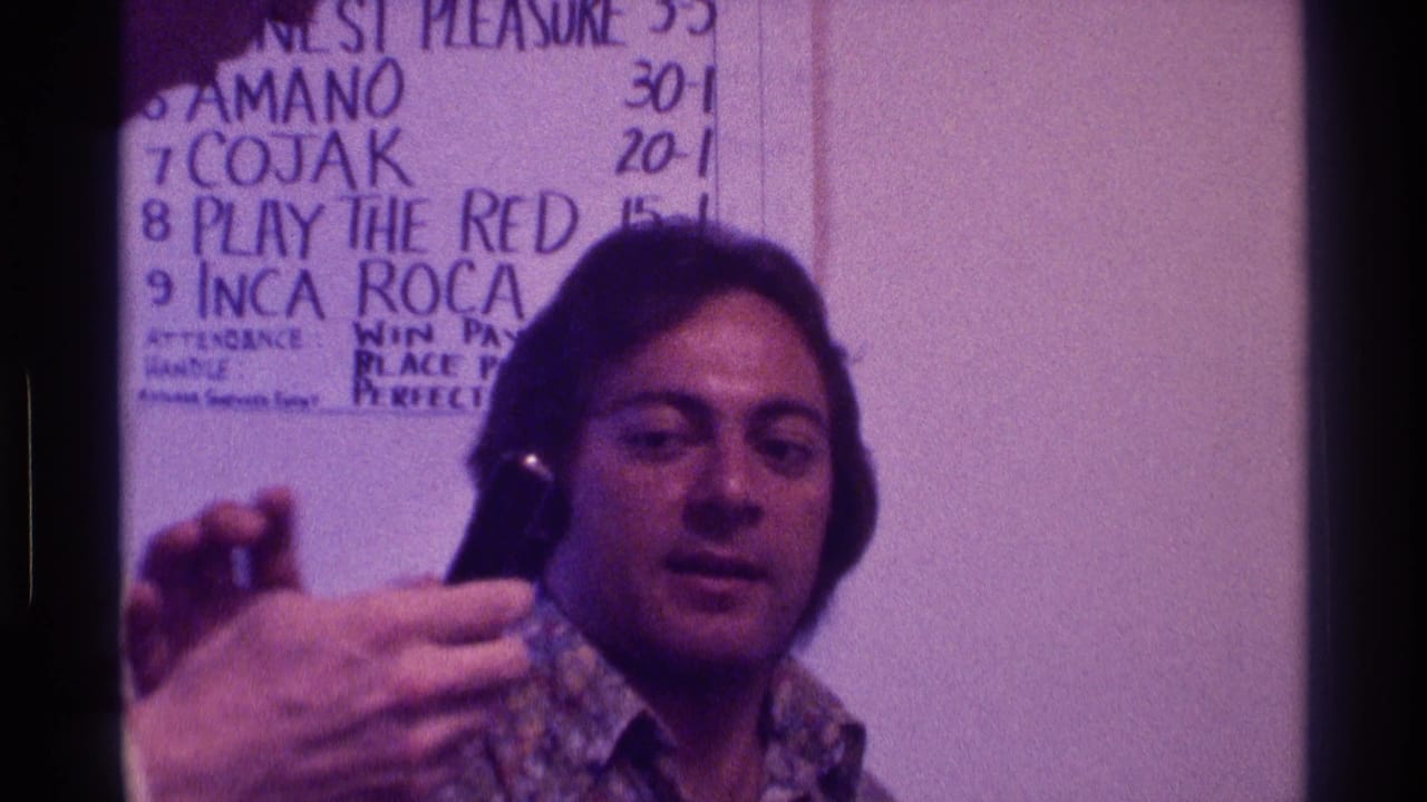 Man in patterned shirt holds a black microphone near a handwritten wall scoreboard.