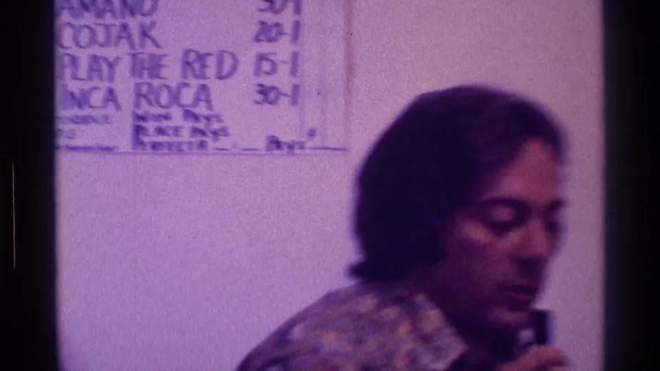 The man turns in profile and appears to speak into a small black microphone, with a handwritten score sheet partly visible behind him.