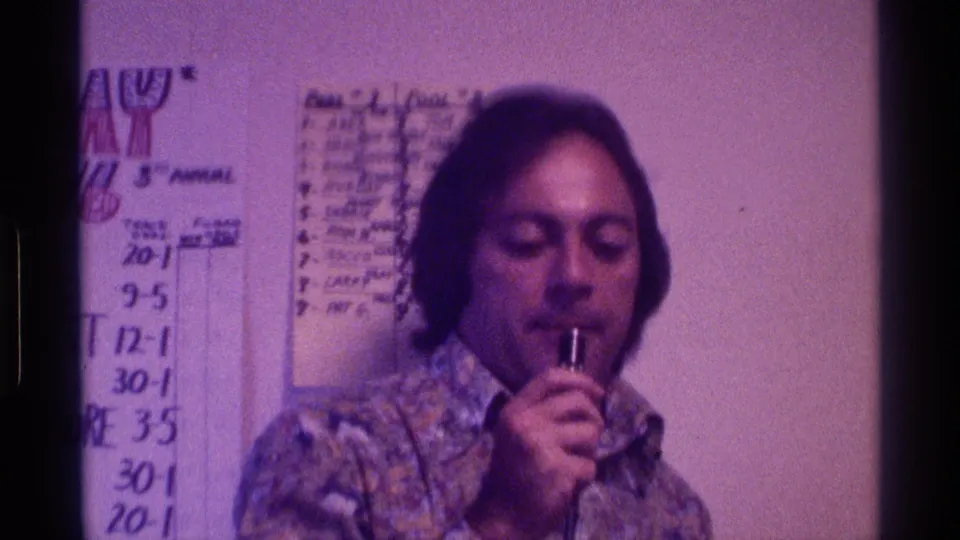 A man in a patterned shirt holds the microphone near his mouth as handwritten charts and score numbers fill the wall behind him.