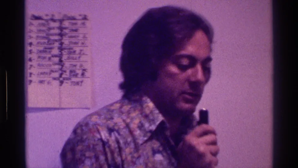 The man looks downward with the microphone in hand, filmed indoors against a pale wall with a handwritten chart in the background.