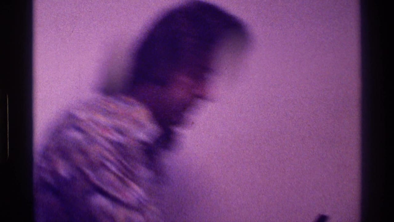 Blurred man in a patterned shirt leaning beside a plain interior wall.