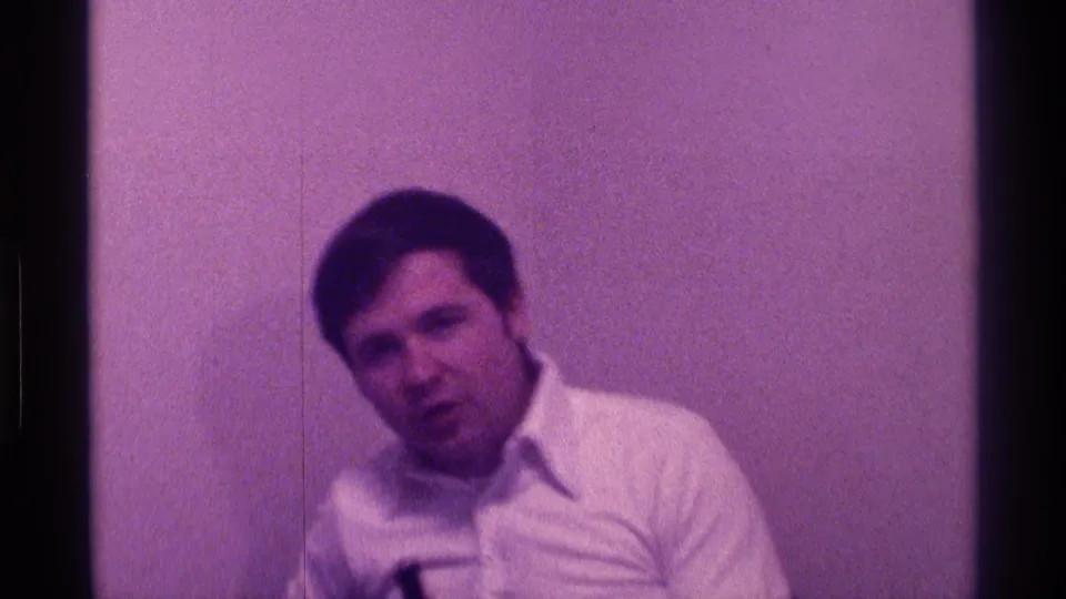 Adult man in a white button-down shirt sits indoors in Kentucky, 1976, holding a black microphone against a plain wall.