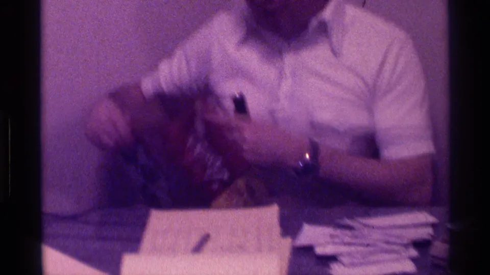 The man in a white shirt sits at a table with papers and a microphone while another adult figure moves across the left side of the frame.