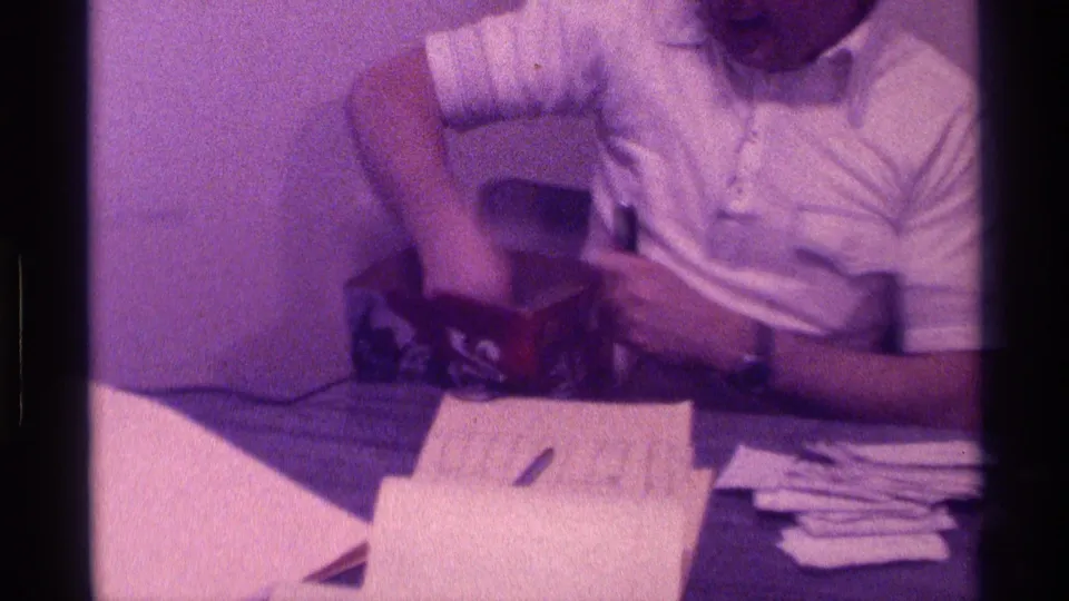 Seated adult man in a white shirt holds a black microphone at an indoor Kentucky event in 1976, with papers, a pen, and a box on the table.