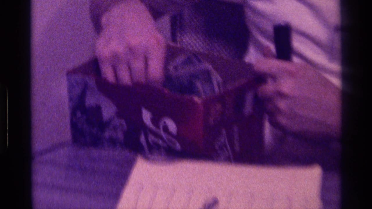 In 1976 Kentucky home-movie footage, an adult man in a white shirt sits indoors with a small box on his lap and papers visible below.