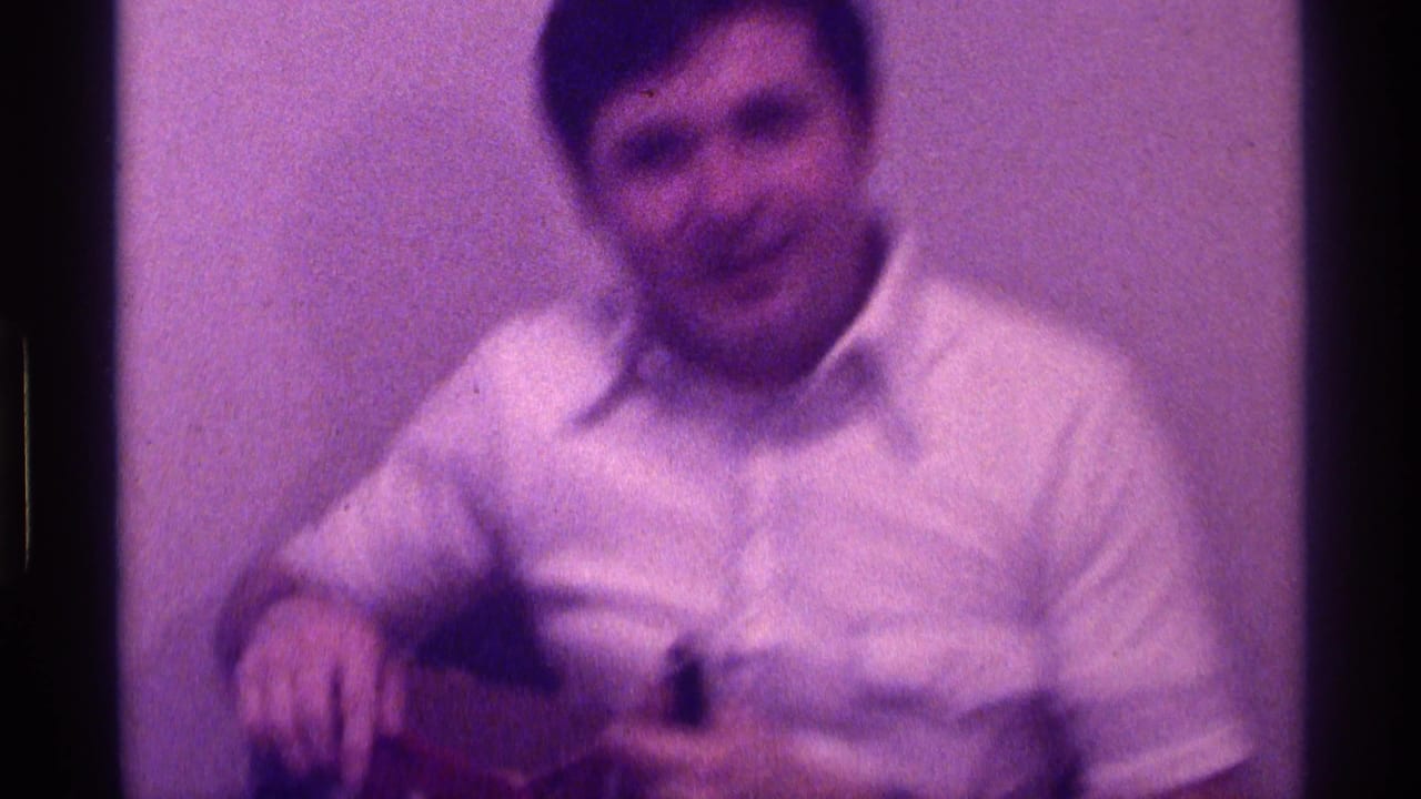 The man sits against a plain wall, smiling toward the camera while holding the box in his lap in the 1976 Kentucky footage.