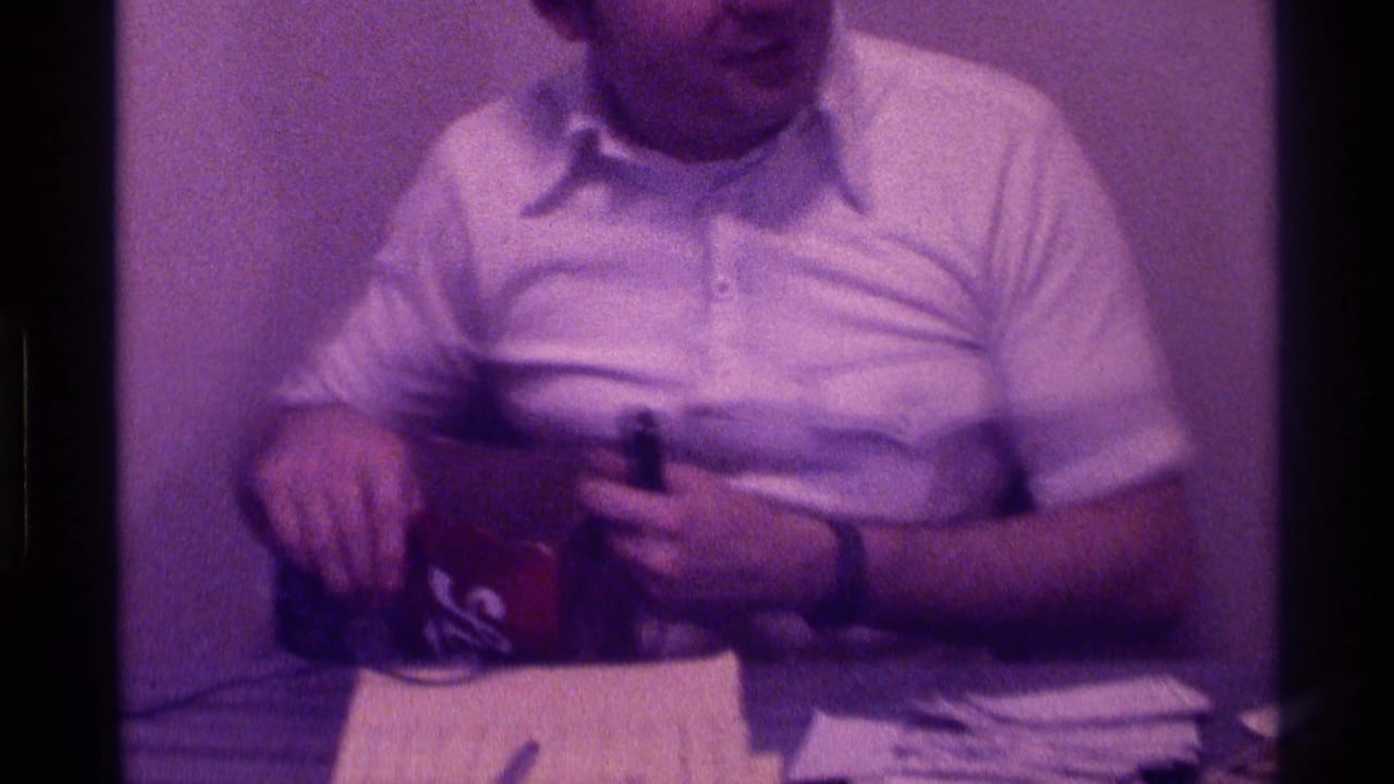 The man looks downward as he handles or points toward the box, with a stack of papers and a pen visible in front of him.