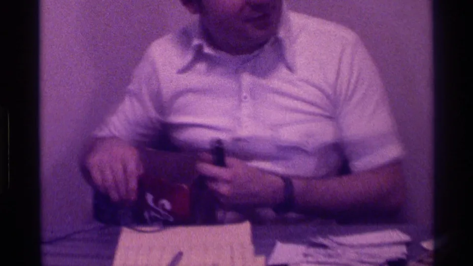 The man looks downward as he handles or points toward the box, with a stack of papers and a pen visible in front of him.