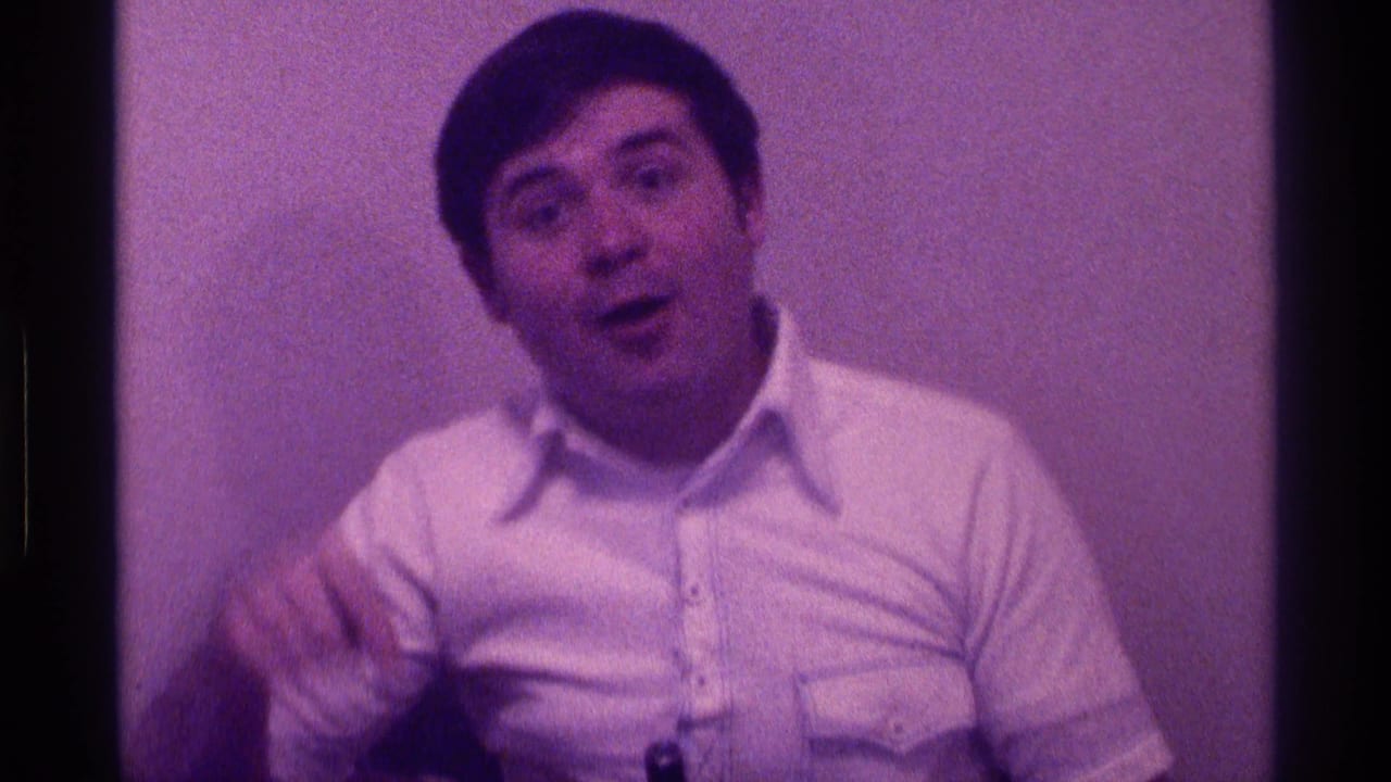 The man faces the camera with an animated expression, appearing to speak while gesturing near the box in a private indoor setting.