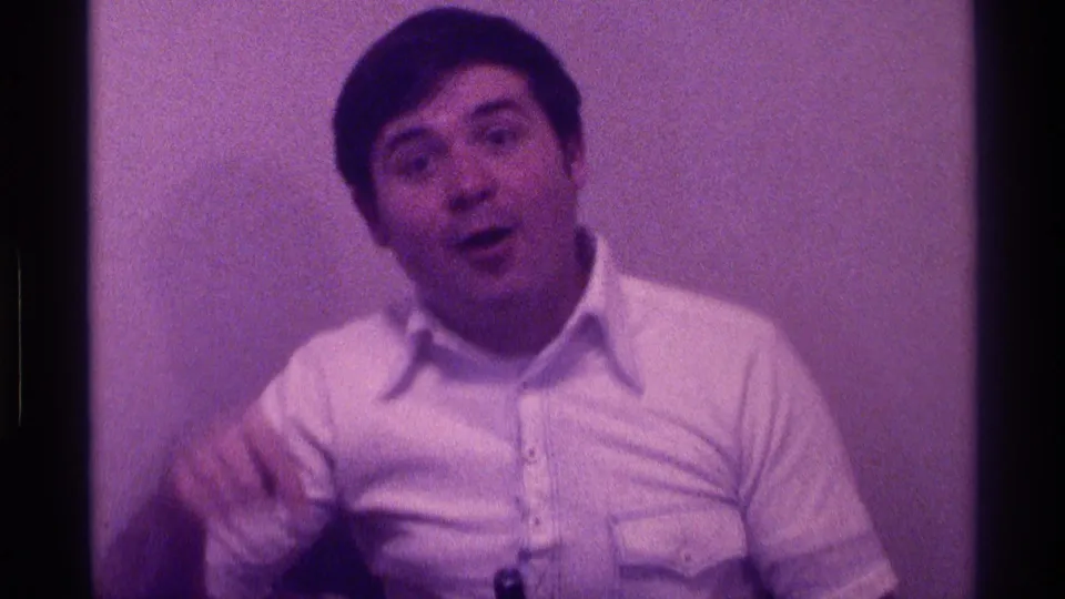 The man faces the camera with an animated expression, appearing to speak while gesturing near the box in a private indoor setting.