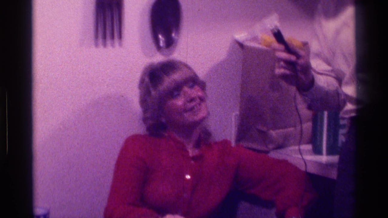In a 1976 Kentucky home-movie frame, a seated woman in a red blouse smiles while an interviewer holds a microphone near her.