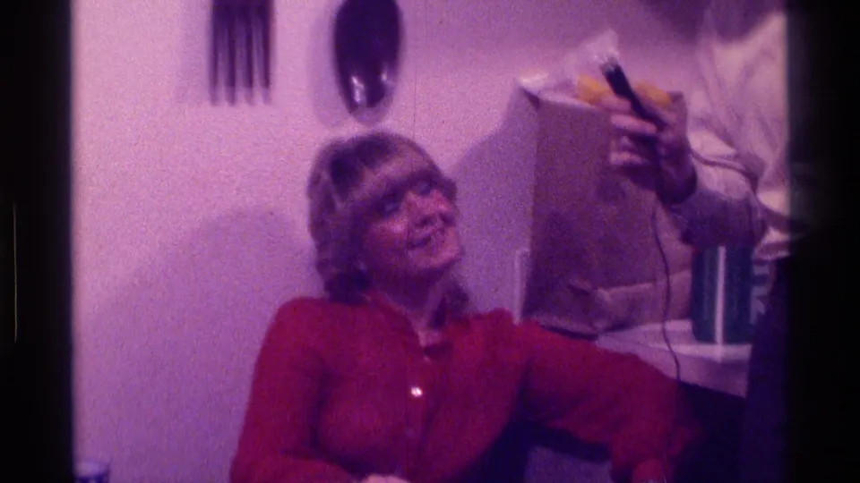 In a 1976 Kentucky home-movie frame, a seated woman in a red blouse smiles while an interviewer holds a microphone near her.