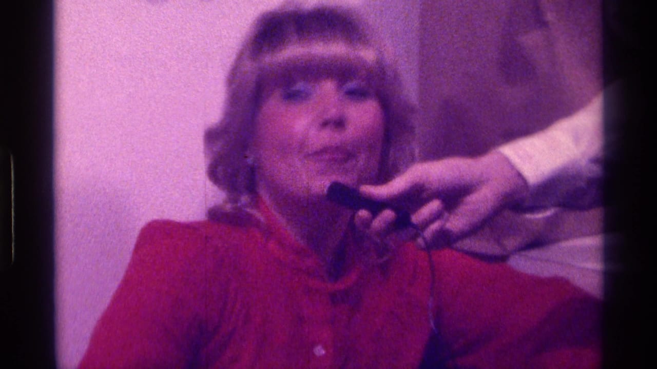 Blonde woman in a red top sings toward a handheld microphone.