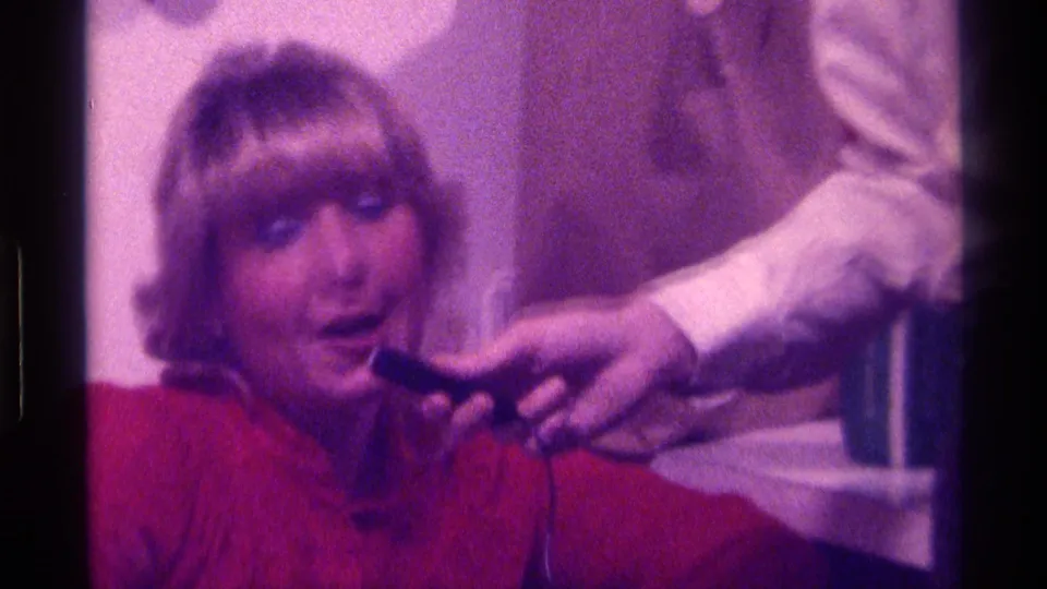 The woman continues singing in the Kentucky interior while a man in a white shirt holds the microphone close.