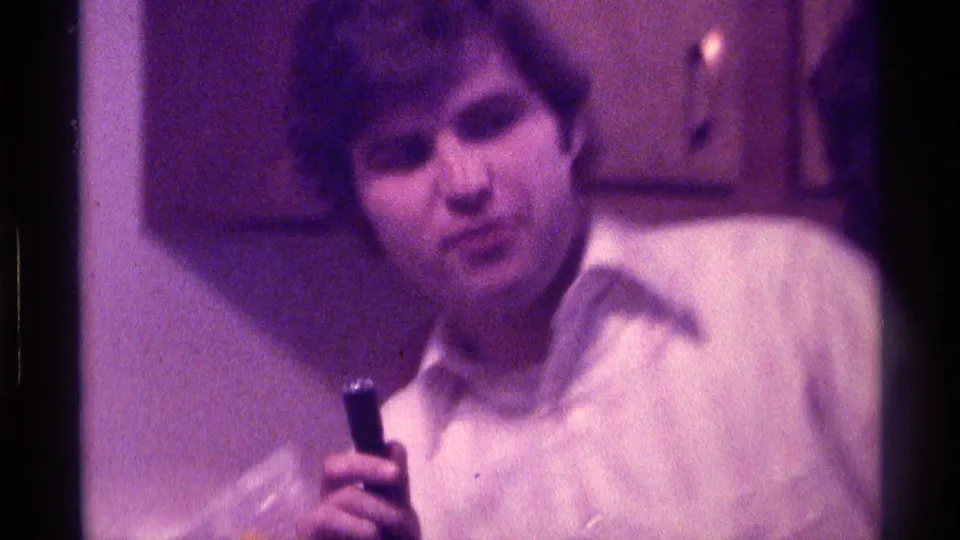 A man in a white shirt appears with the handheld microphone inside the same Kentucky home-movie interior.