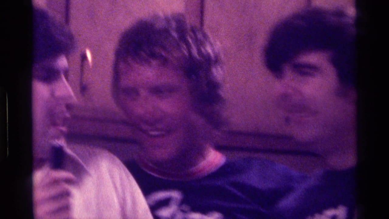 Three adult men stand close together in a kitchen, laughing and leaning into one another.