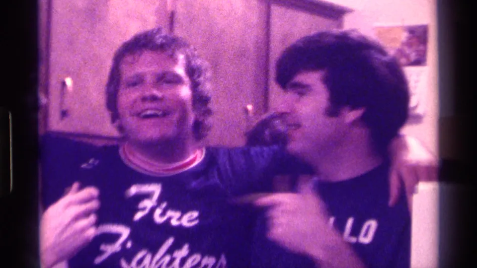 Two adult men in a Kentucky kitchen stand with arms around each other, wearing readable slogan shirts and gesturing toward the camera.