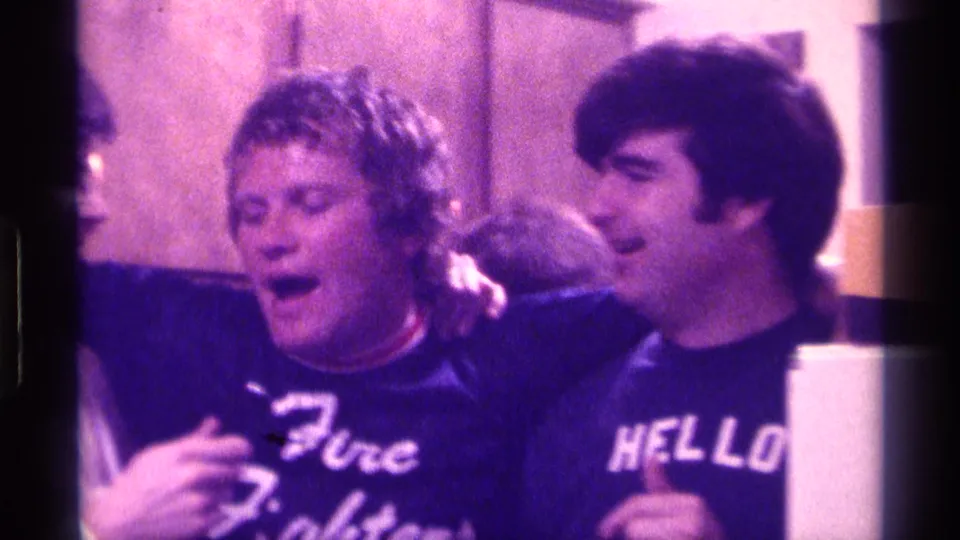 Two adult men in a 1970s Kentucky kitchen lean together, with one reacting animatedly while wearing a Fire Fighters shirt.