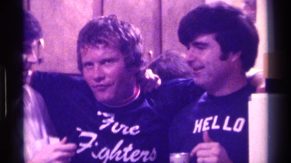 Two adult men in a 1976 Kentucky kitchen stand arm in arm, smiling near the camera with cups visible below.