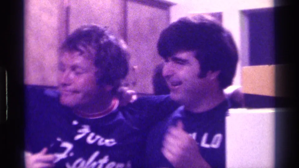 Two young men in 1970s casual shirts smile beside a visible microphone, with indoor wall panels and a pale cabinet behind them.