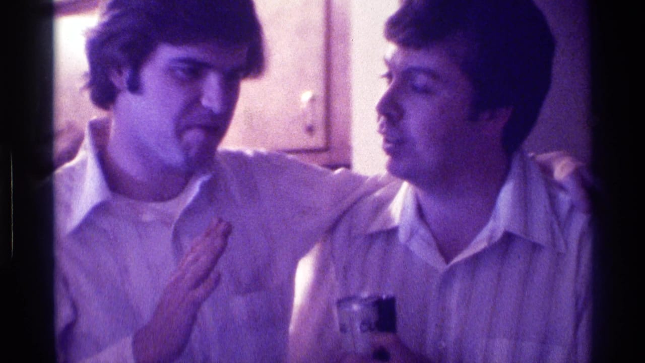 Two men in collared shirts sit close together indoors, one with an arm around the other.