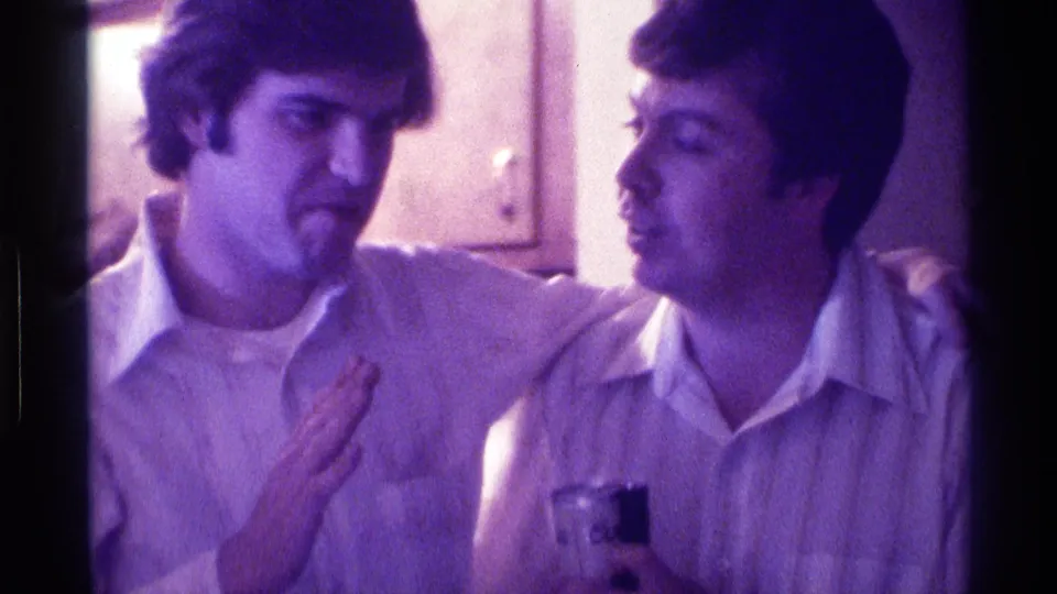 1976 Kentucky home-movie frame of two young men in collared shirts seated close together at an indoor gathering, one holding a drink.