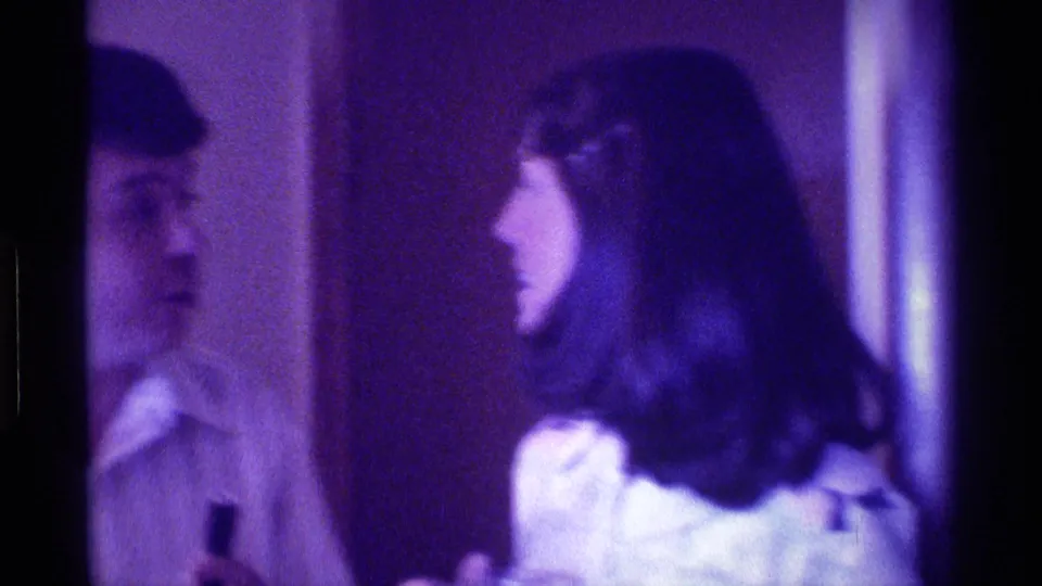 1976 Kentucky home-movie frame showing a man and dark-haired woman talking in a dim private interior with purple color cast.